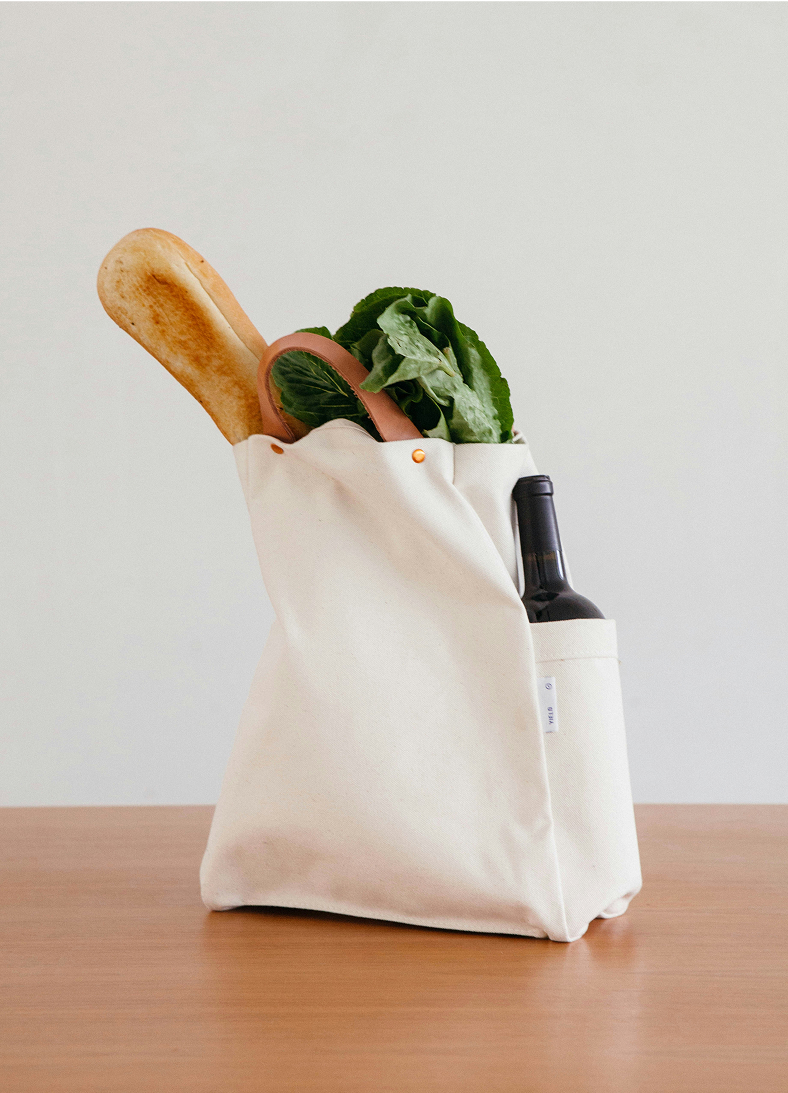 A white reusable tote bag filled with a baguette, lettuce, and a bottle of wine on a wooden surface