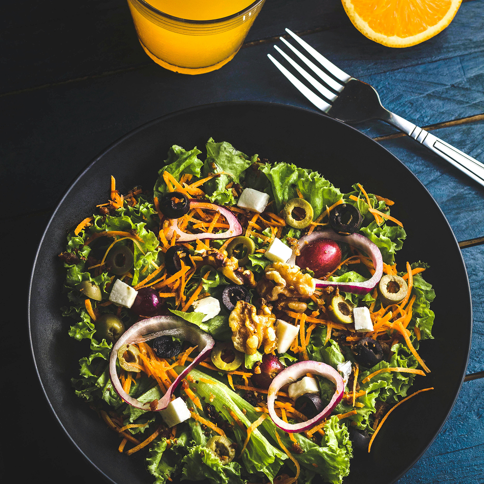 A black bowl filled with a colorful mixed salad, an orange half, a fork, and a glass of orange juice on a dark blue surface