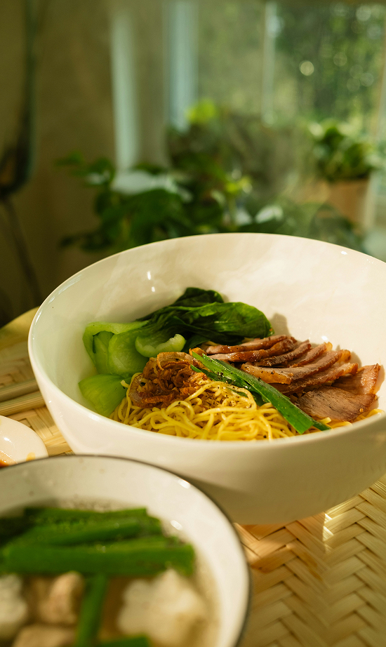 A white bowl filled with egg noodles, slices of roasted duck, and green leafy vegetables