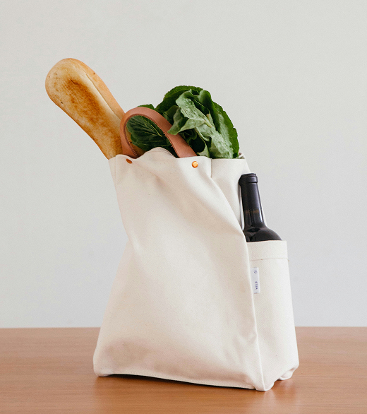 A white reusable tote bag filled with a baguette, lettuce, and a bottle of wine on a wooden surface