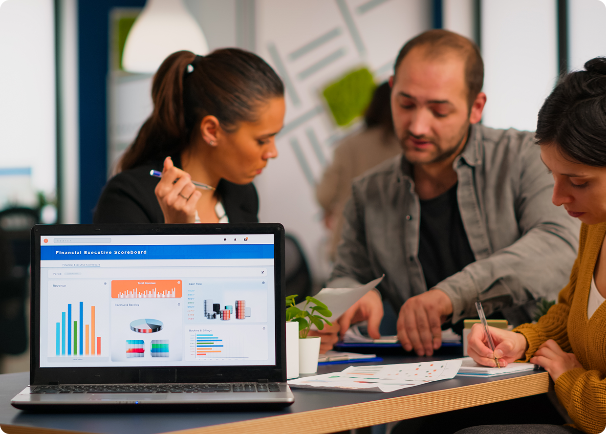 Three professionals collaborate around a table, reviewing financial data displayed on a laptop screen, with two taking notes.
